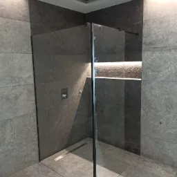 Nowoczesna kabina prysznicowa walk-in z czarnymi profilami, szklaną ścianką, odpływem liniowym i ciemnoszarą okładziną kamienną na ścianach oraz podłodze, z podświetlaną wnęką.