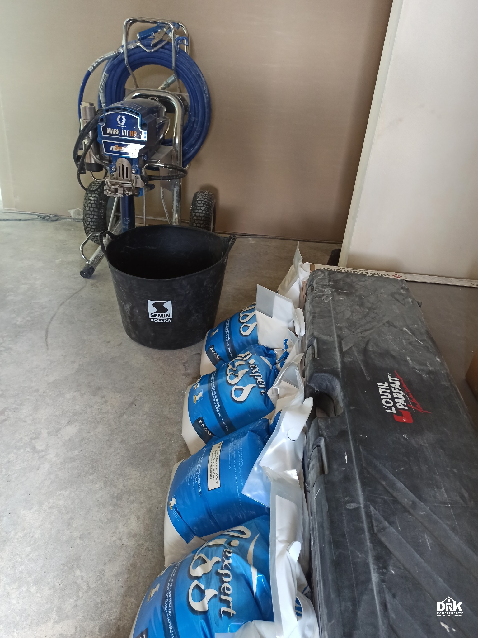 Agregat malarski Graco Mark VII, czarne wiadro z logo Semin Polska, kilka worków z białym proszkiem Expert oraz czarna skrzynia na narzędzia L'Outil Parfait stoją na betonowej podłodze przy ścianie.