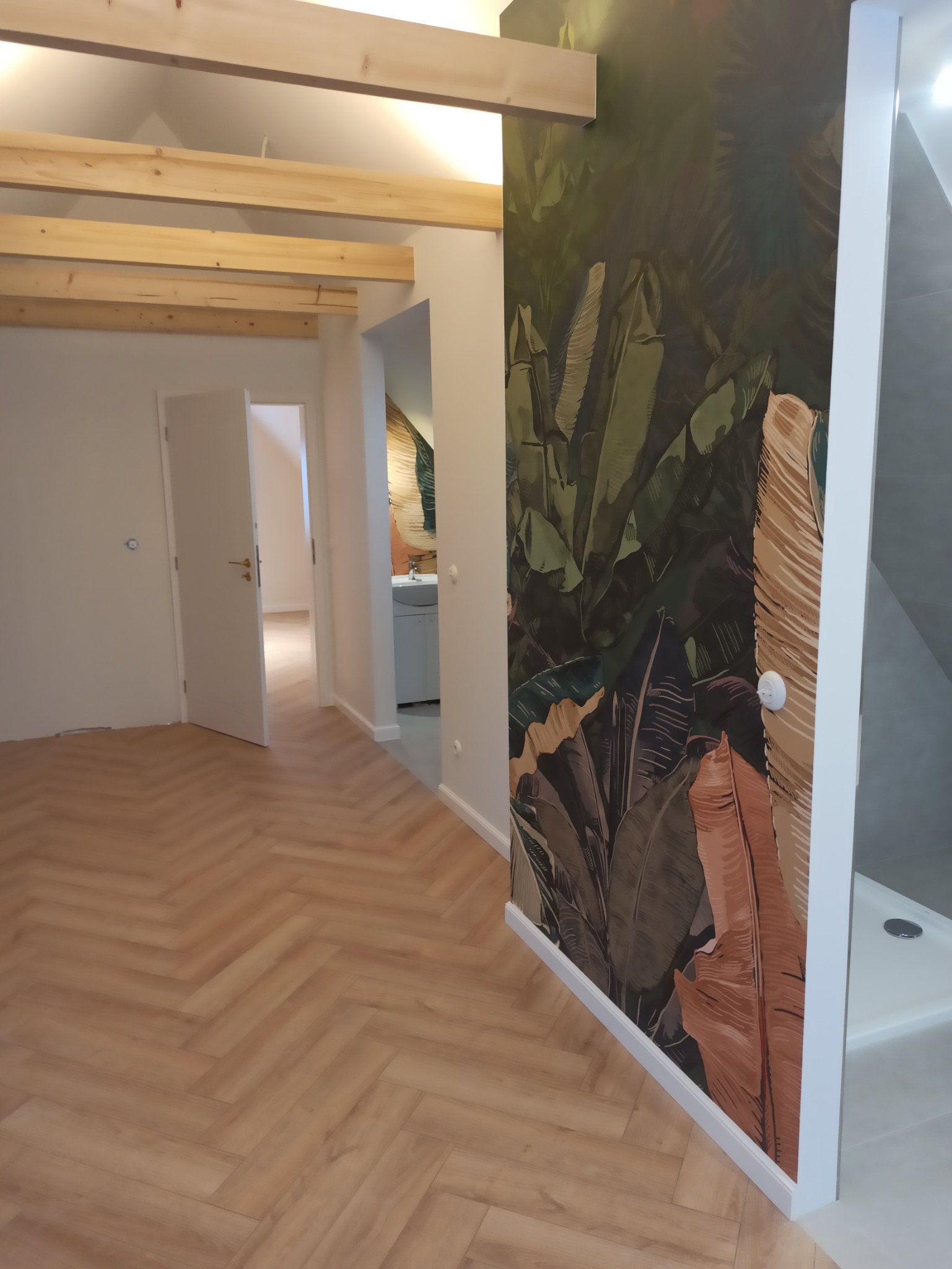 Wnętrze po remoncie: parkiet ułożony w jodełkę, ściana z motywem tropikalnych liści, drewniane belki pod sufitem, widok na łazienkę z kabiną prysznicową.