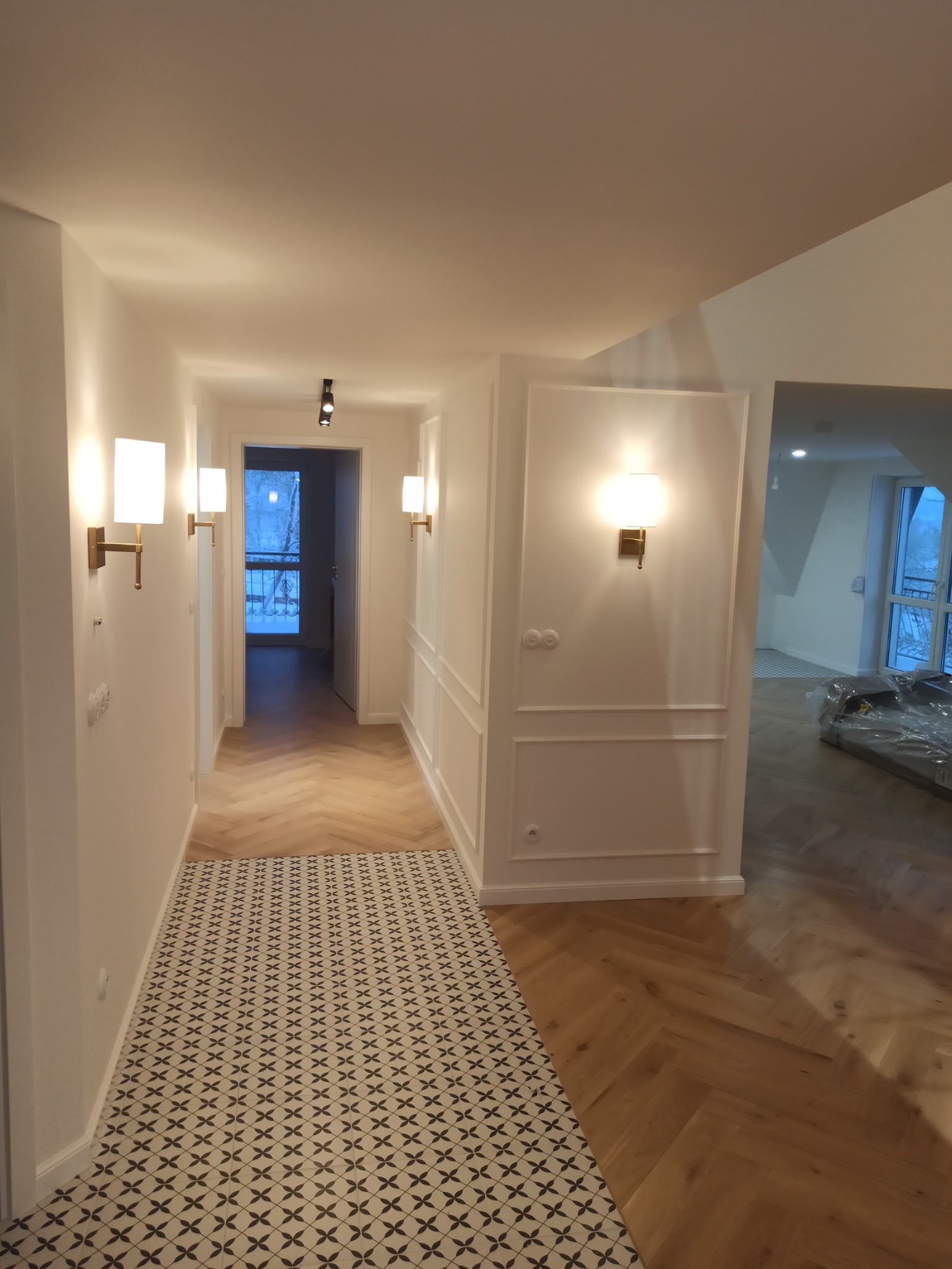 Korytarz po remoncie w Jabłonce z panelami podłogowymi ułożonymi w jodełkę, fragmentem podłogi z wzorzystych płytek, białymi ścianami z dekoracyjnymi listwami i oświetleniem ściennym.