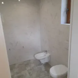 Jasna łazienka z marmurowymi płytkami na ścianach, bidetem i toaletą, szare płytki podłogowe, okno z drewnianą ramą.