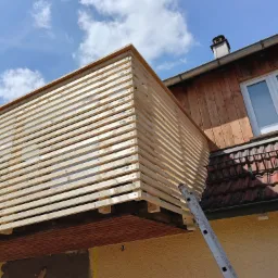 Nowo wybudowany, drewniany balkon z poziomymi listwami, widok z dołu na tle błękitnego nieba z chmurami, drabina oparta o ścianę budynku z drewnianą elewacją i czerwonym dachem.