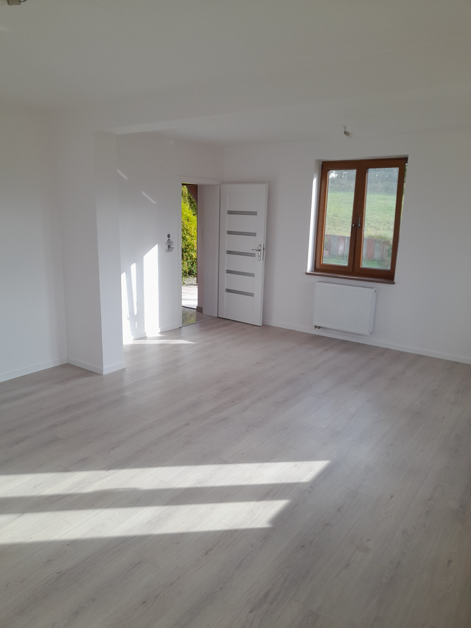 Jasne, puste pomieszczenie z panelami podłogowymi w odcieniach szarości, białymi ścianami, drewnianym oknem z widokiem na zieleń i białą, nowoczesną, otwartą stolarką drzwiową.