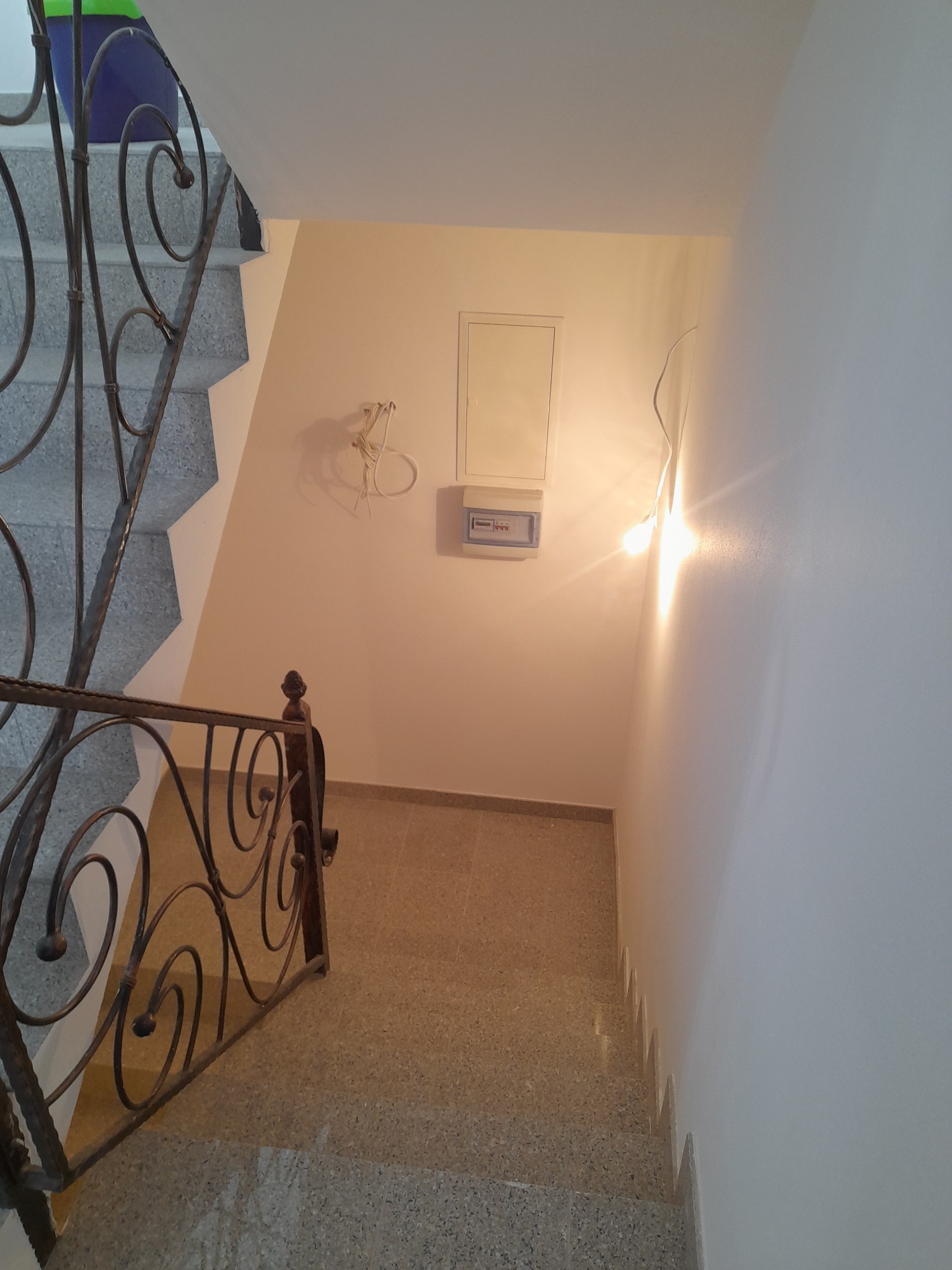 Klatka schodowa z ozdobną, metalową balustradą, widok z góry na dół, na ścianie widać białą skrzynkę elektryczną i wystające kable, ściany świeżo pomalowane na biało.