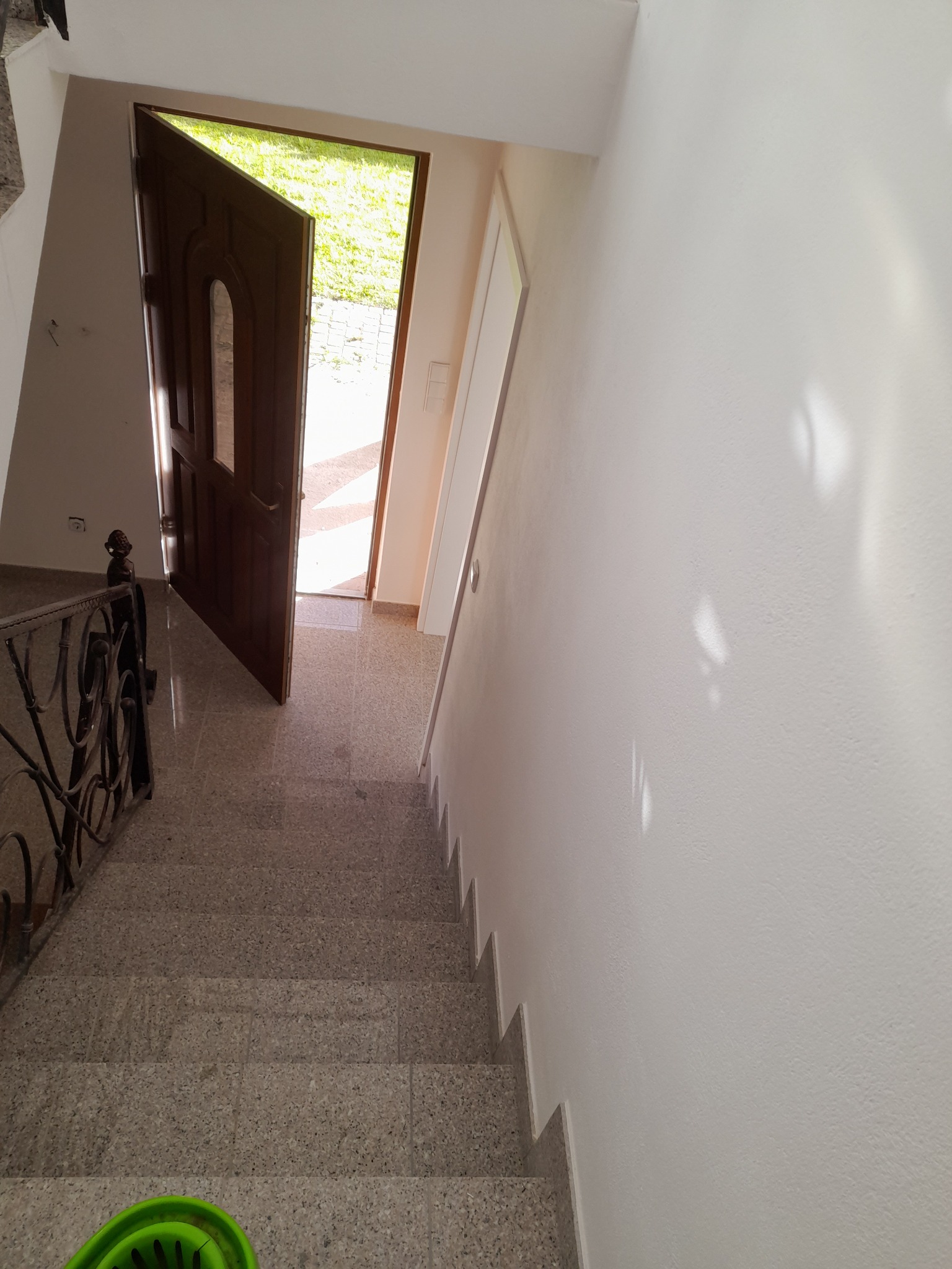 Widok z góry na klatkę schodową z terazzo, z otwartymi drzwiami wejściowymi w tle i fragmentem ozdobnej, żelaznej balustrady po lewej stronie; świeżo pomalowana, biała ściana po prawej.