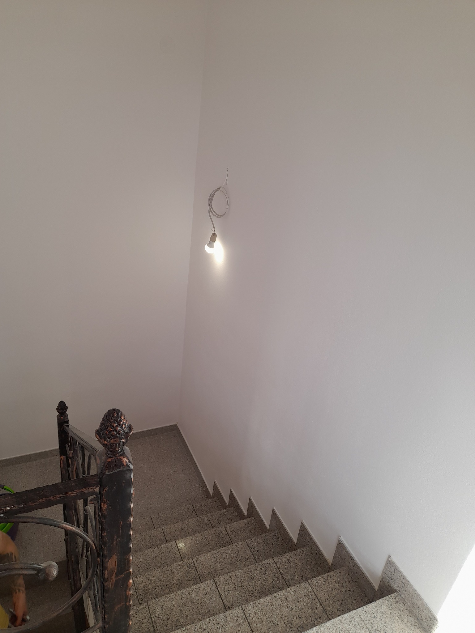 Świeżo pomalowana klatka schodowa z szarymi płytkami na stopniach, widok z góry na metalową balustradę i żarówkę na kablu, po remoncie.