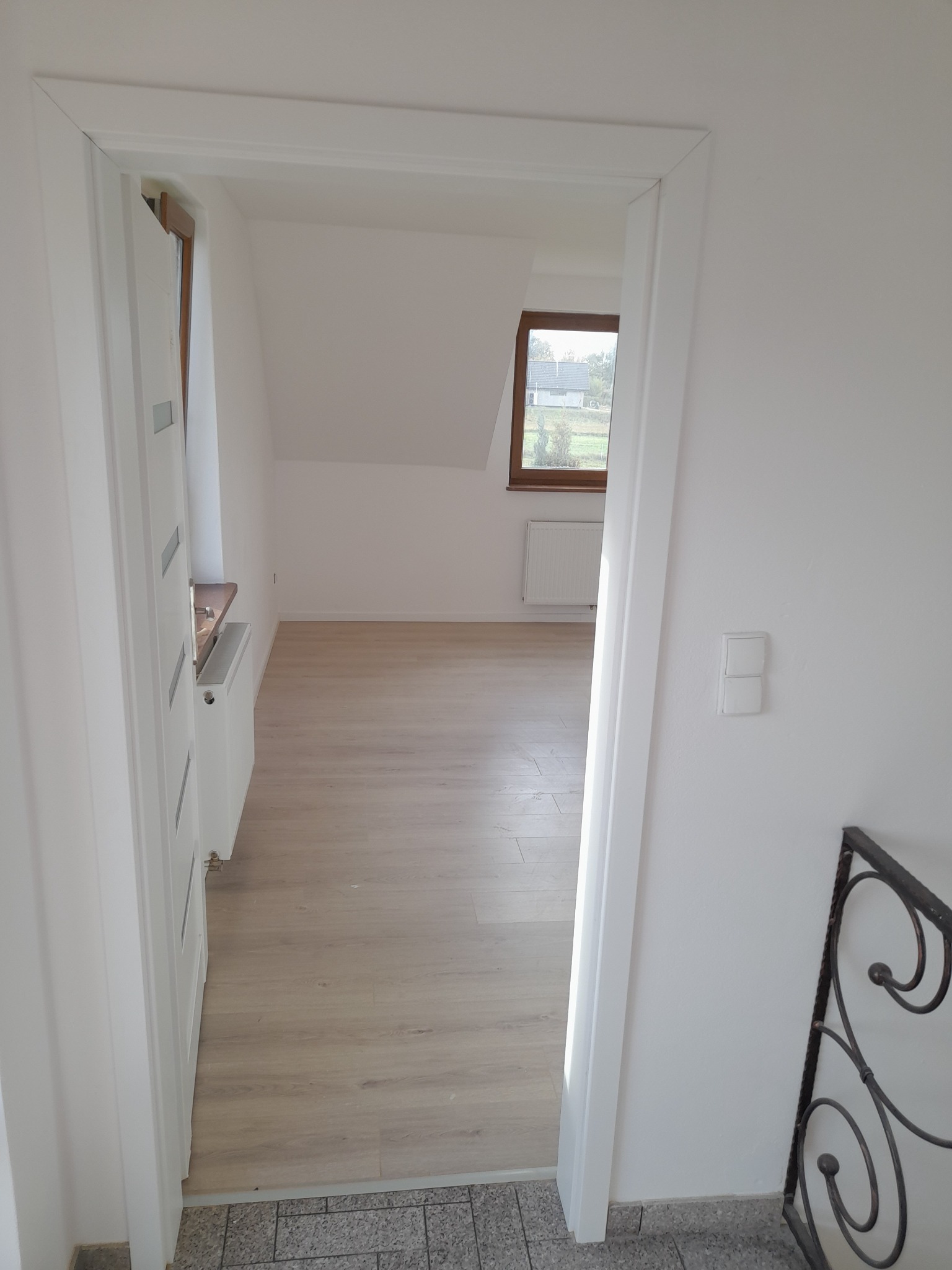 Jasne panele podłogowe w nowo wykończonym pokoju na poddaszu, widoczne przez białe futryny drzwi, z fragmentem grzejnika i okna dachowego w tle.