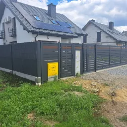 Nowoczesne ogrodzenie panelowe w kolorze grafitowym z poziomymi elementami drewnianymi, skrzynka gazowa żółta, dom jednorodzinny z panelami słonecznymi w tle, trawa i ziemia na pierwszym planie.