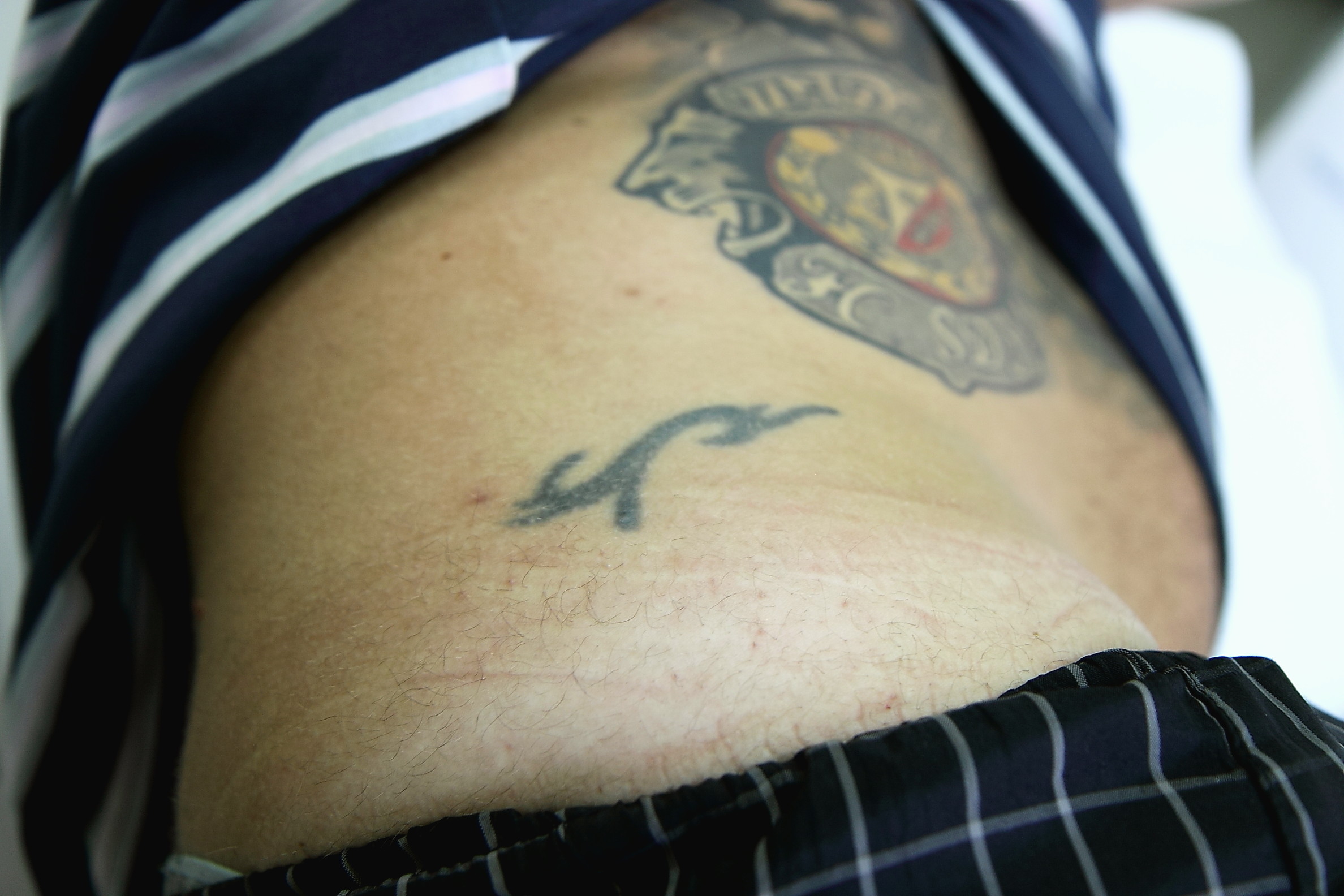 Fragment ciała z dwoma tatuażami: większym kolorowym na górze i mniejszym czarnym poniżej, widoczny fragment ubrania w paski i kratę.