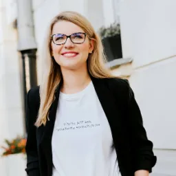 Uśmiechnięta blondynka w okularach i czarnej marynarce, ubrana w białą koszulkę z napisem 'Fajnie jest być świadomą swoich praw', pozuje na tle jasnego budynku.