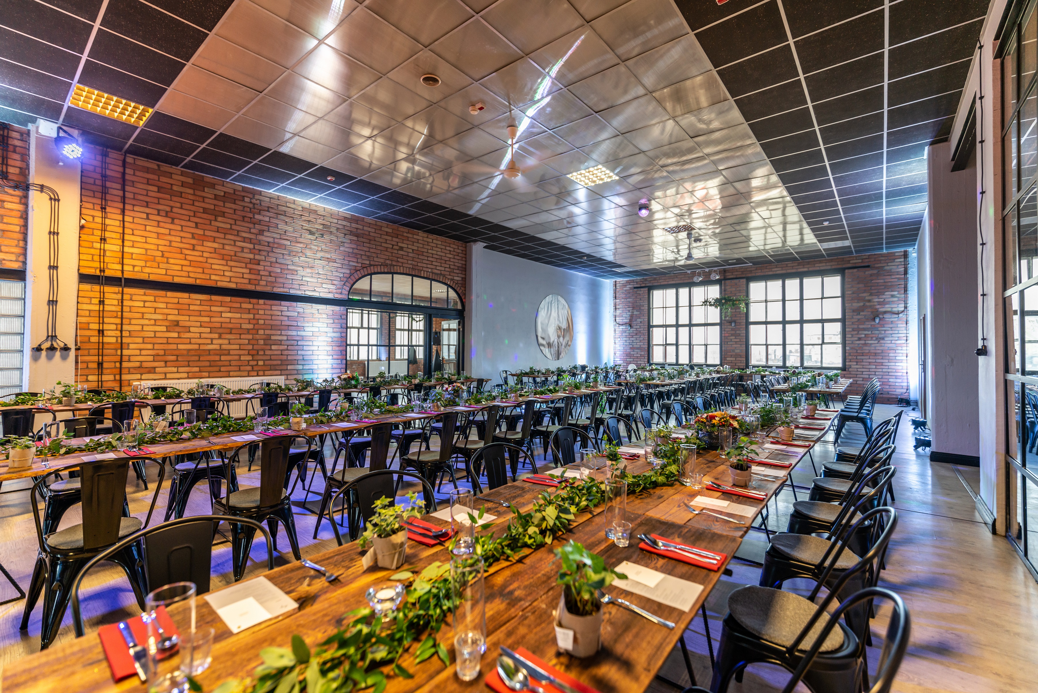 Uroczysta sala z długimi drewnianymi stołami, czarnymi krzesłami i dekoracjami z zieleni. Czerwone serwetki i srebrne sztućce na stołach. Ceglana ściana i nowoczesne oświetlenie.