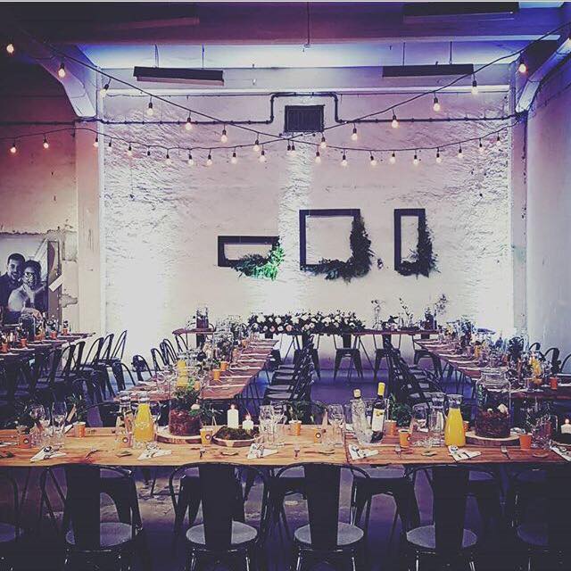 Industrialna sala weselna z długimi drewnianymi stołami, czarnymi krzesłami, dekoracją z żarówek i kompozycjami kwiatowymi w geometrycznych ramach na białej ceglanej ścianie.