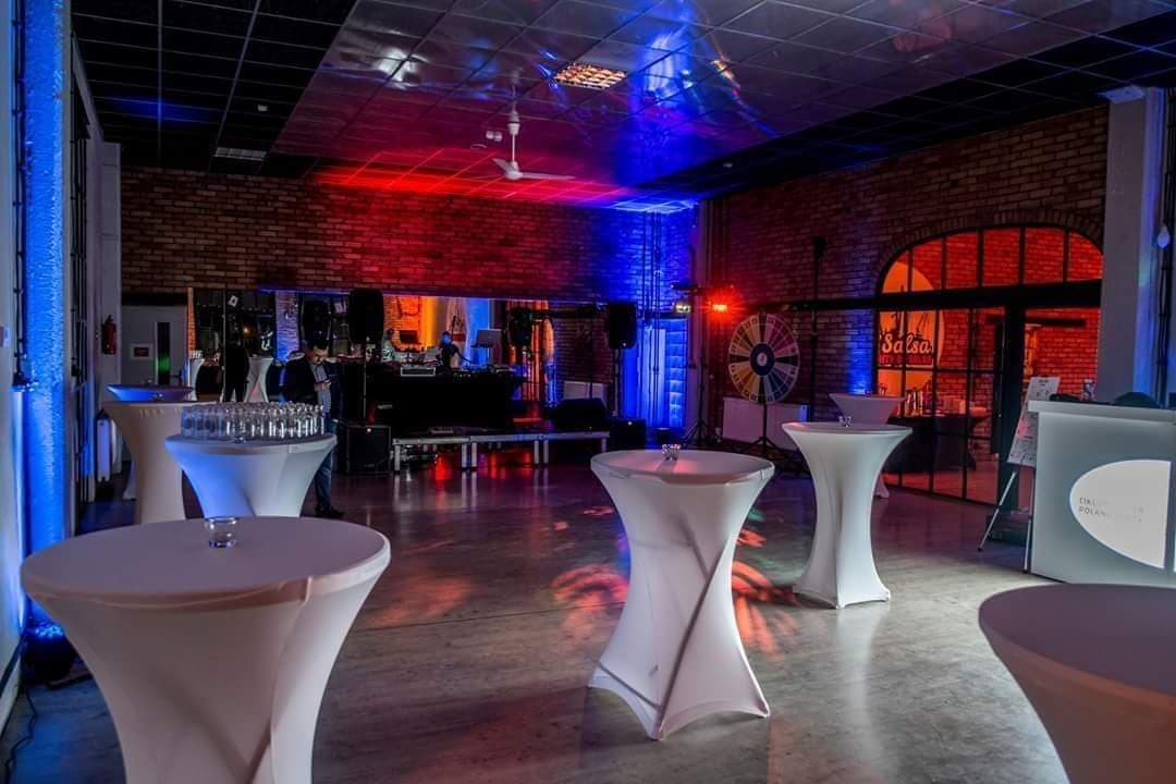 Sala eventowa z dekoracją świetlną w kolorach czerwieni i błękitu, wysokie stoliki koktajlowe z białymi pokrowcami, DJ na scenie w tle, ceglane ściany.