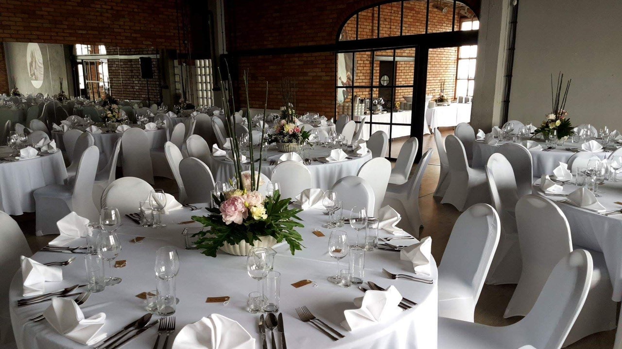 Uroczysta sala bankietowa z okrągłymi stołami przykrytymi białymi obrusami, nakryciem i dekoracjami kwiatowymi; krzesła w białych pokrowcach, ceglane ściany w tle.