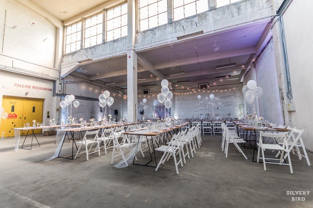 Uroczysta sala w stylu industrialnym z długimi stołami, białymi krzesłami, balonami i dekoracjami kwiatowymi.