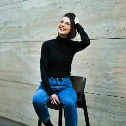 Uśmiechnięta kobieta w okularach siedzi na krześle, dotykając włosów na tle ściany z trawertynu. Spojrzenie skierowane w górę.