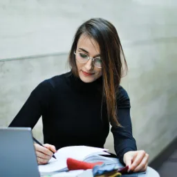 Projektantka wnętrz w czarnym golfie, z okularami, analizuje próbki tkanin, notując w notatniku. Widoczny laptop i jasne tło.