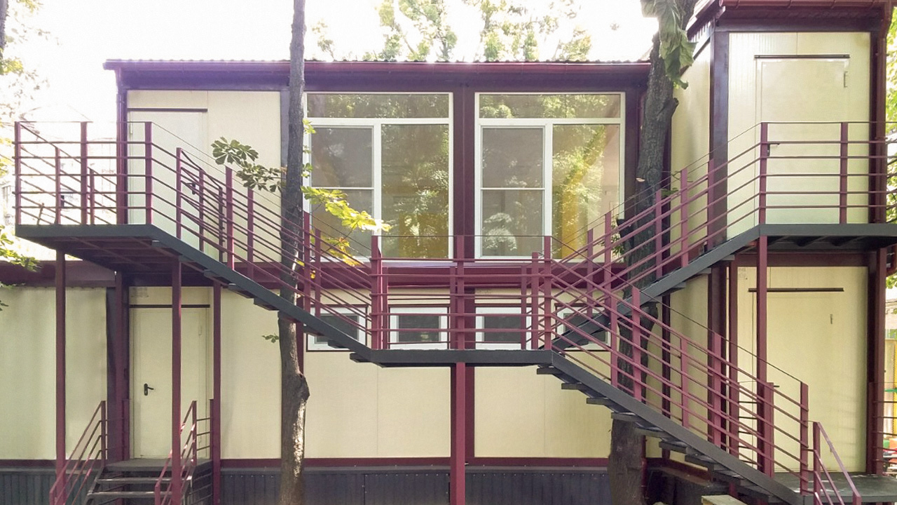 Dwukondygnacyjny budynek z beżowymi ścianami i bordowymi elementami konstrukcyjnymi, schody zewnętrzne z metalową balustradą, drzewa wkomponowane w konstrukcję.