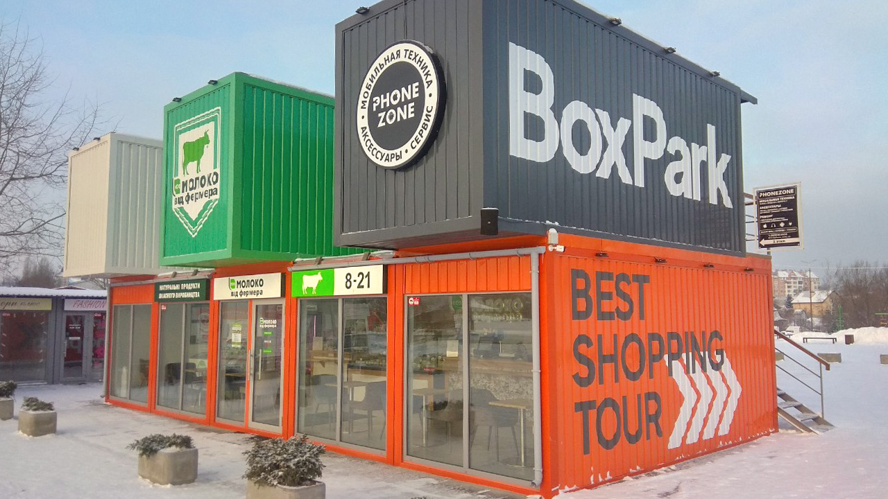 Zewnętrzny widok pawilonów handlowych zbudowanych z kontenerów, z szyldami 'BoxPark', 'Phone Zone' i 'Mleko od farmera'.
