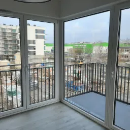 Widok z balkonu na plac budowy i sklep Leroy Merlin w tle, przez duże okna z białymi ramami. Szare panele podłogowe w pomieszczeniu.