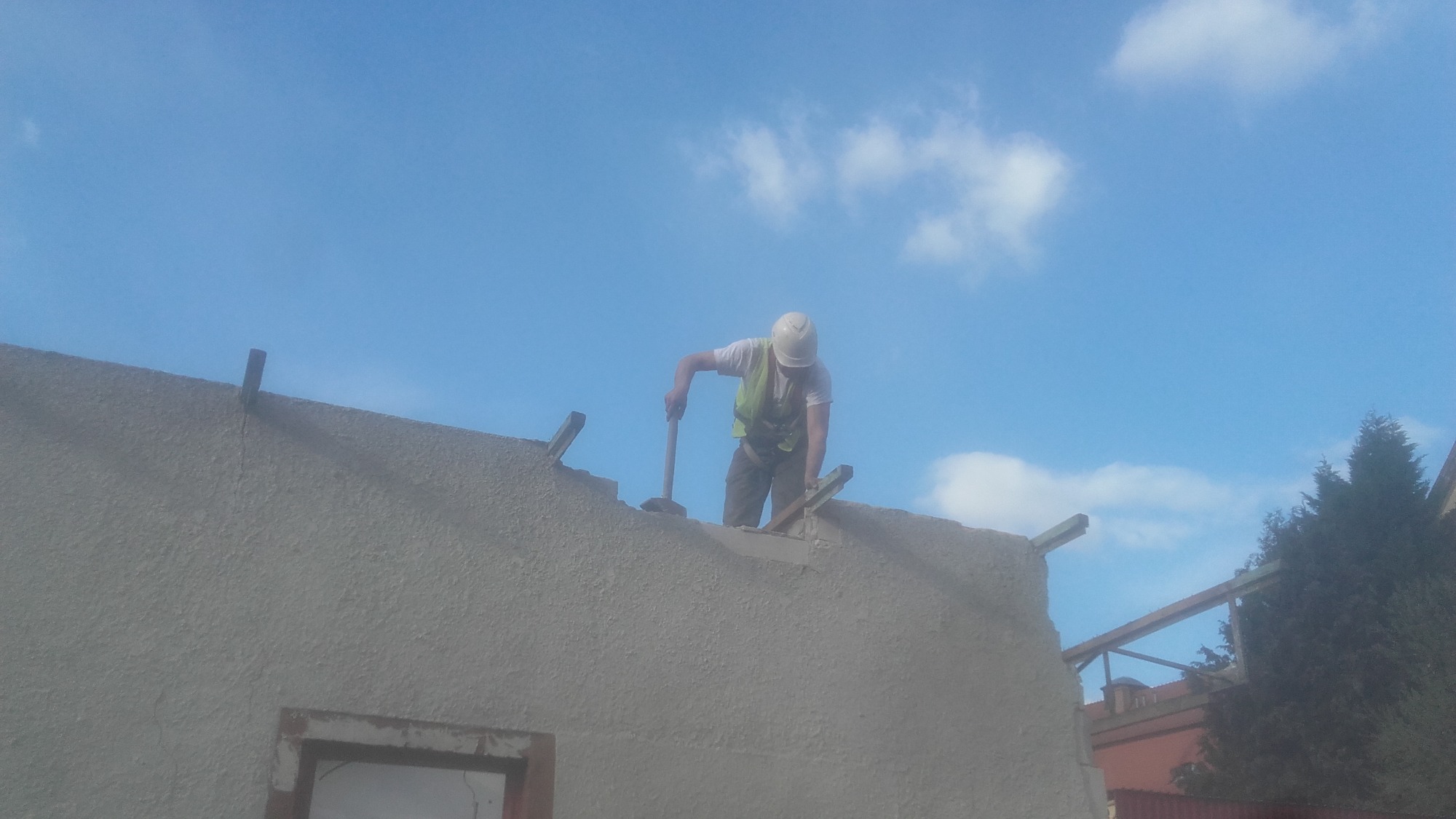 Pracownik w kasku i kamizelce uderza młotem w mur, prawdopodobnie podczas prac rozbiórkowych lub demontażowych, na tle błękitnego nieba z chmurami.