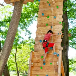 Chłopiec w czerwonej koszulce w paski wspina się po drewnianej ściance wspinaczkowej w parku linowym, zabezpieczony liną i uprzężą.