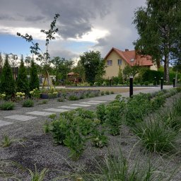 PRODECO BEATA BACH - Nowoczesny ogród przydomowy z geometrycznymi ścieżkami z płyt, ozdobnymi trawami i rabatami, na tle domu z czerwoną dachówką i pochmurnego nieba.