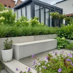 Nowoczesny ogród z betonową ławką, trawami ozdobnymi i kwitnącymi geranium. W tle przeszklona oranżeria i budynki mieszkalne.