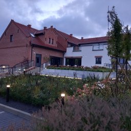 PRODECO BEATA BACH - Ogród z oświetleniem przy budynku z czerwonej cegły i białymi elementami. Widoczne rabaty z różnymi gatunkami roślin i traw ozdobnych, oświetlone lampami.