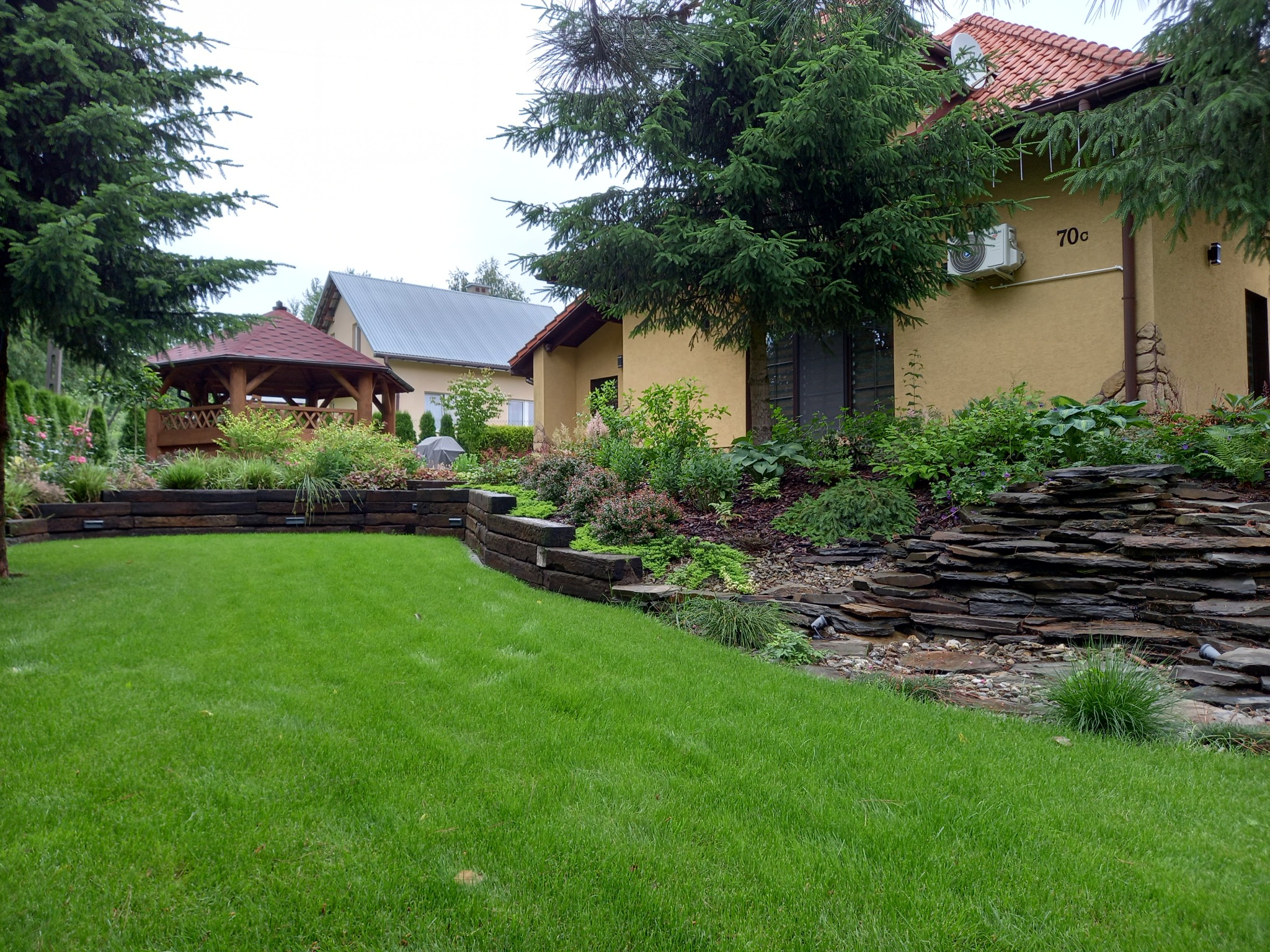 Zadbany ogród z równo przystrzyżonym trawnikiem, drewnianą altaną i kaskadą z kamieni. Dom z kremową elewacją i klimatyzatorem w tle.