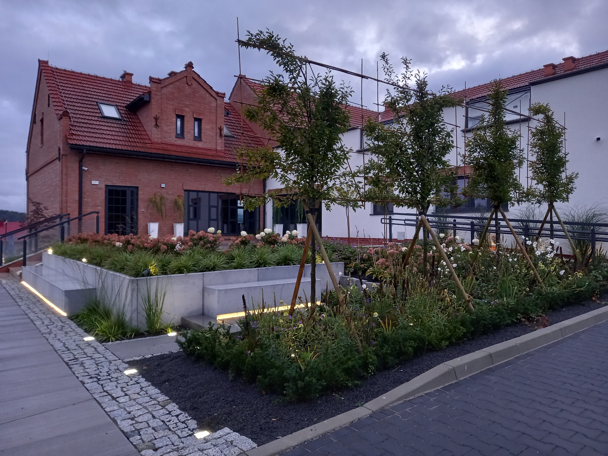 Nowoczesny ogród z podświetlonymi rabatami i młodymi drzewkami na tle budynku z czerwonej cegły i białego budynku. Oświetlony chodnik z kostki.