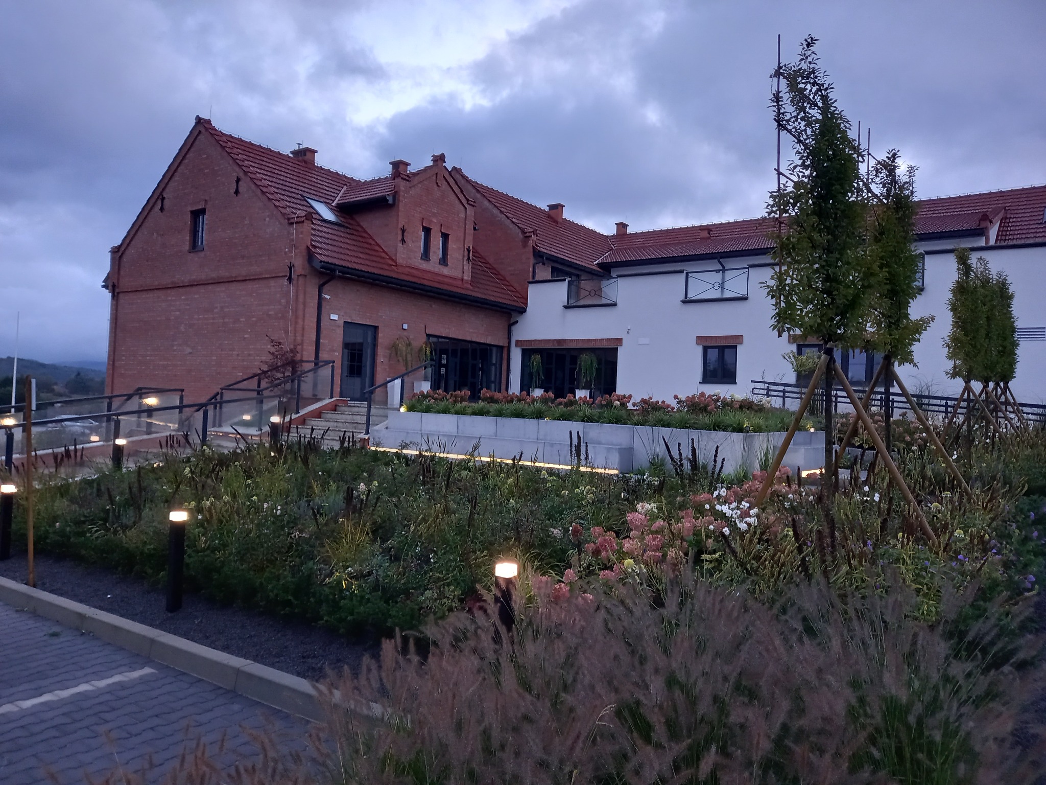 Ogród z oświetleniem przy budynku z czerwonej cegły i białymi elementami. Widoczne rabaty z różnymi gatunkami roślin i traw ozdobnych, oświetlone lampami.