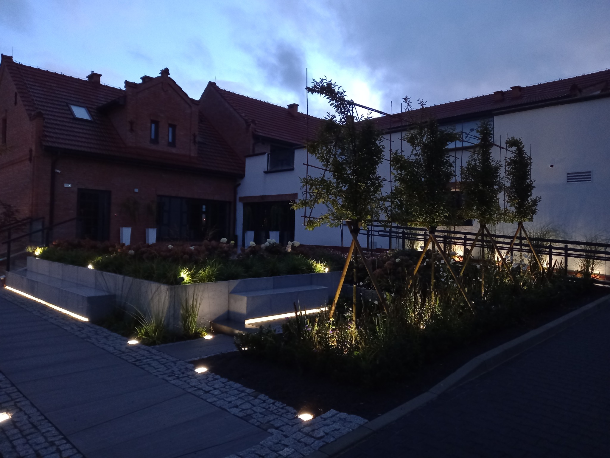 Oświetlony ogród przy posesji w wieczornym klimacie. Podświetlone rośliny i ścieżki tworzą unikalny nastrój. Architektura budynku w tle.