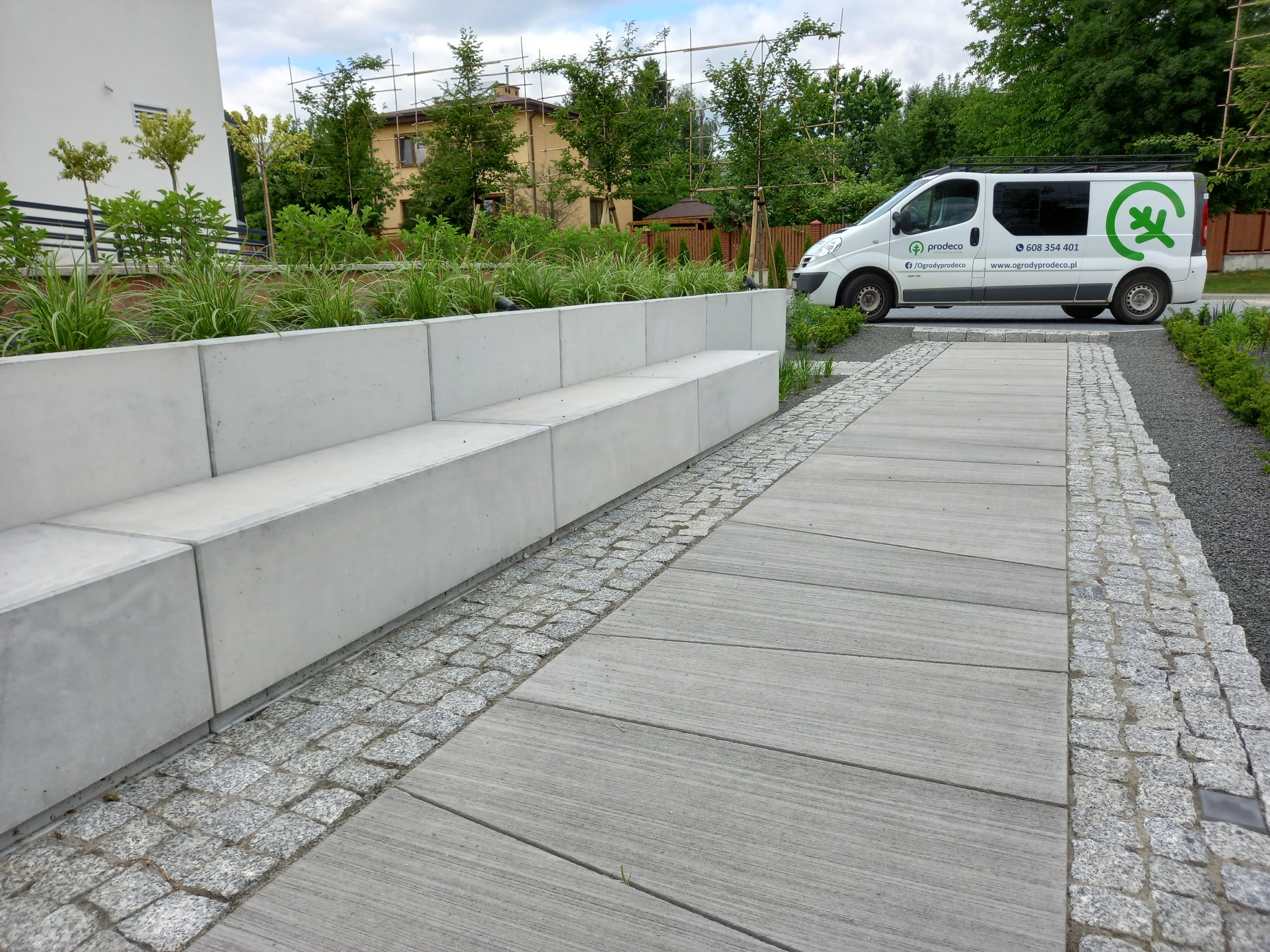 Nowoczesny ogród z betonową ławką i ścieżką z płyt, obok zaparkowany biały samochód firmy ogrodniczej. Widoczne drzewa i krzewy.