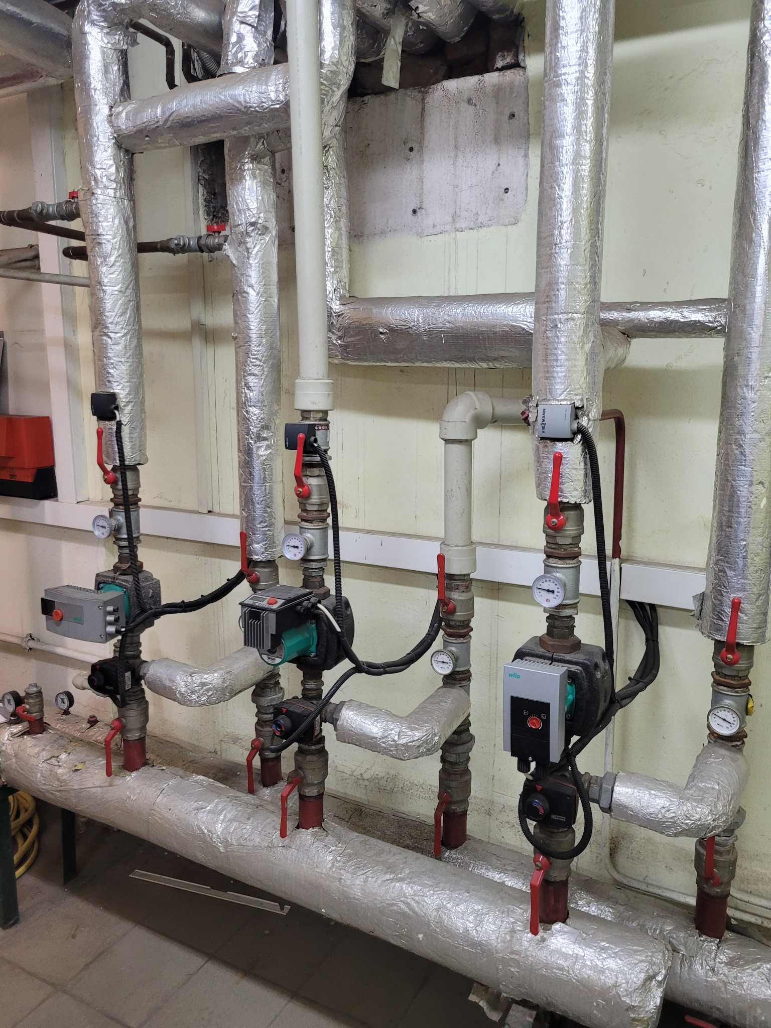 Izolowane rury grzewcze z pompami obiegowymi i zaworami, widoczne manometry i elementy sterujące, instalacja w pomieszczeniu technicznym.