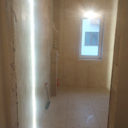 Krzoch-Bud - Wnętrze łazienki w trakcie remontu, widoczne płytki na ścianach i podłodze, niezakończone prace wykończeniowe przy ościeżnicy i wystające przewody elektryczne.