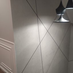 Krzoch-Bud - Szpachlowana ściana w odcieniach szarości z geometrycznym wzorem z czarnych linii, oświetlona designerską lampą wiszącą i sąsiadująca z białą ścianą z dekoracyjnymi panelami.