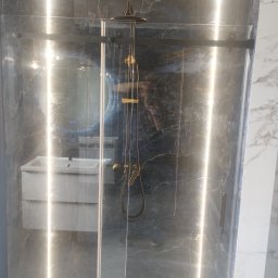 Krzoch-Bud - Szklana kabina prysznicowa z deszczownicą w łazience wykończonej marmurem. Widoczne oświetlenie LED i umywalka z szafką.