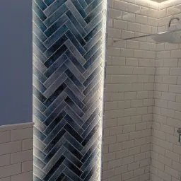 Ściana w łazience z niebieską mozaiką ułożoną w jodełkę, podświetloną listwą LED. Obok białe, prostokątne płytki i fragment kabiny prysznicowej.