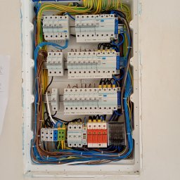 Krzoch-Bud - Rozdzielnica elektryczna Hager w białej obudowie, z widocznymi bezpiecznikami, przewodami w różnych kolorach i listwami zaciskowymi, umieszczona w ścianie w trakcie montażu.