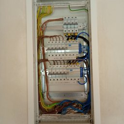 Krzoch-Bud - Szczegółowe ujęcie rozdzielnicy elektrycznej w białej obudowie, z widocznymi kablami w kolorach: niebieskim, brązowym, żółto-zielonym, oraz zabezpieczeniami.