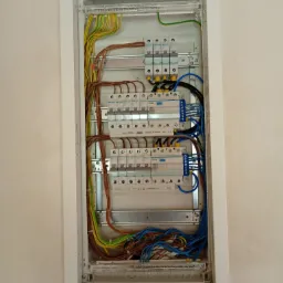 Szczegółowe ujęcie rozdzielnicy elektrycznej w białej obudowie, z widocznymi kablami w kolorach: niebieskim, brązowym, żółto-zielonym, oraz zabezpieczeniami.