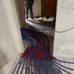 Krzoch-Bud - Rozłożone rury ogrzewania podłogowego w kolorze niebieskim i czerwonym na folii z kratką, widoczne obliczenia na ścianie i fragment osoby w tle.