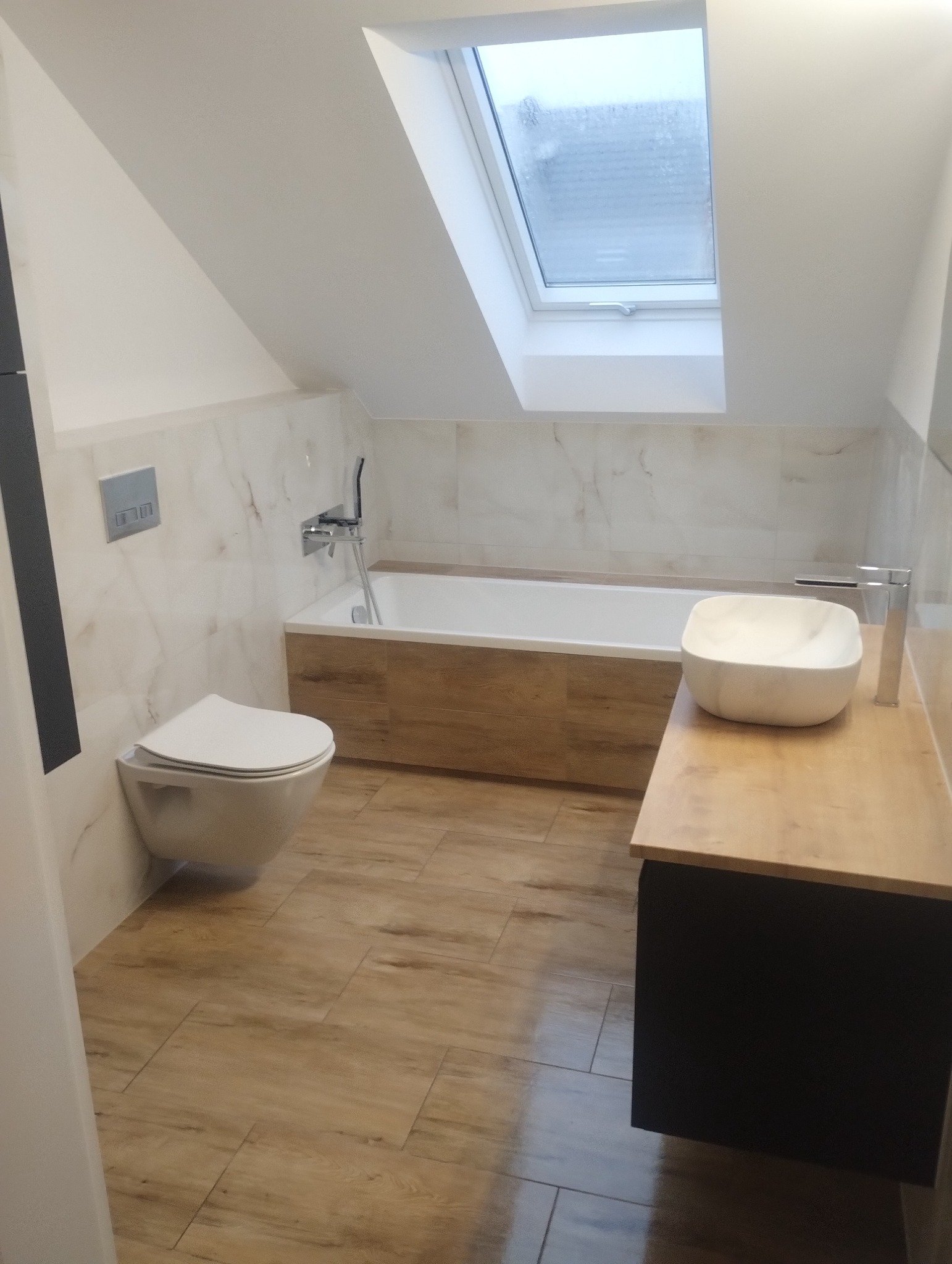 Wykończona łazienka na poddaszu w Stargardzie z wanną obudowaną drewnem, umywalką nablatową na drewnianym blacie i podwieszanym WC, oświetlona przez okno dachowe.