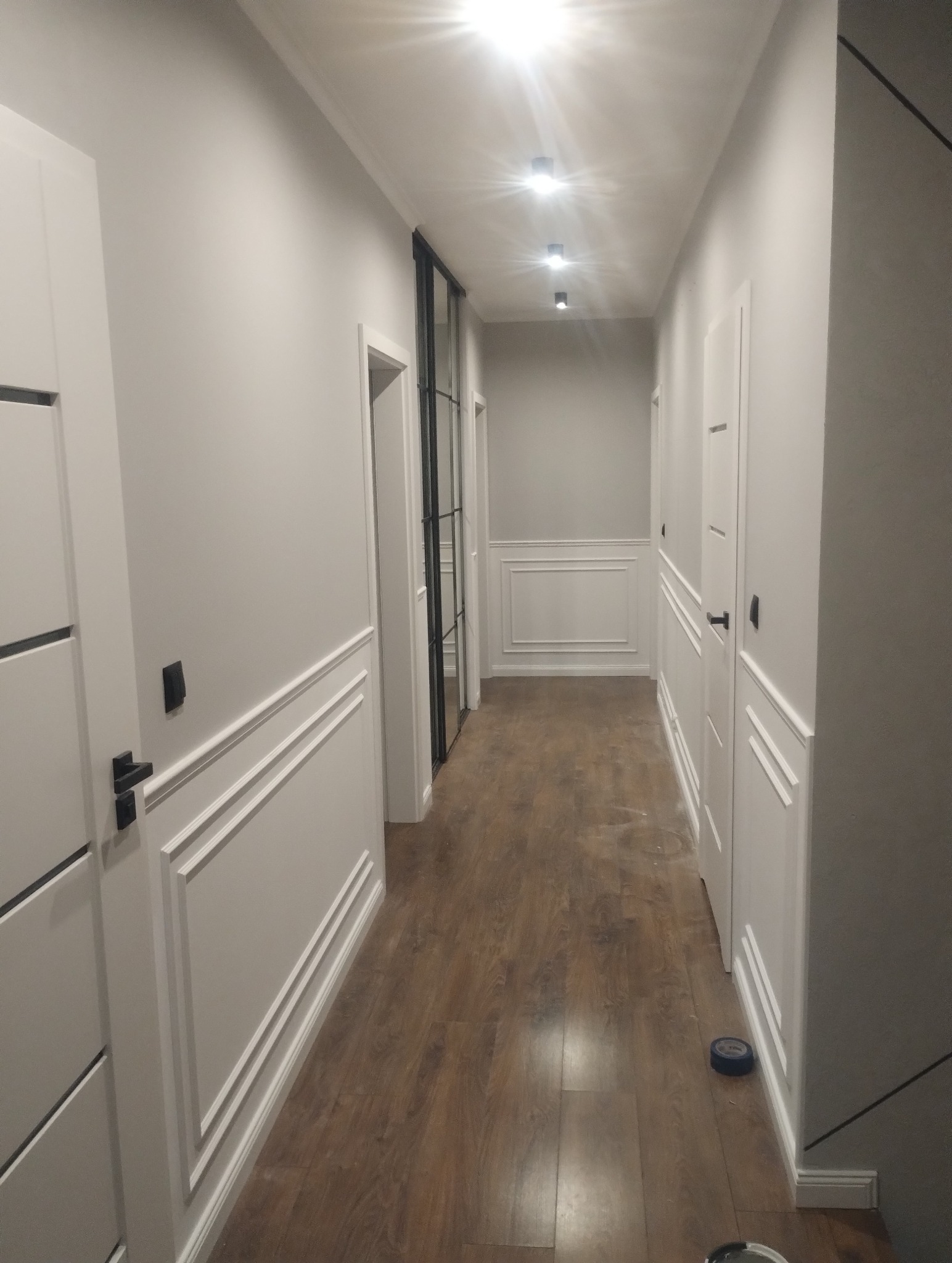 Korytarz po remoncie z drewnopodobną podłogą, białymi ścianami z dekoracyjnymi listwami, czarnymi detalami i punktowym oświetleniem sufitowym.