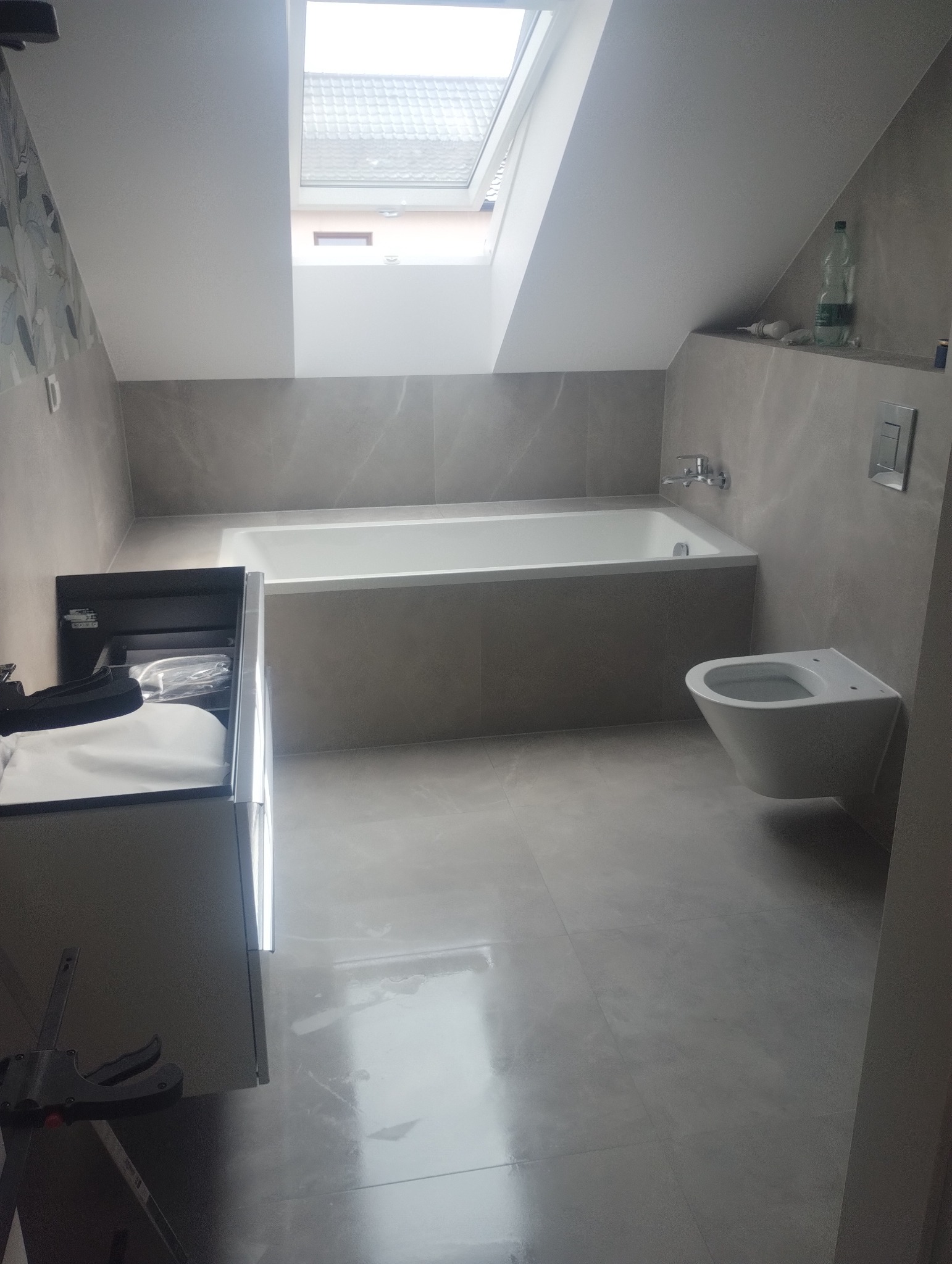 Nowoczesna łazienka na poddaszu z oknem dachowym, szarą armaturą i minimalistycznym designem. Widoczna wanna, toaleta podwieszana i umywalka z szafką.