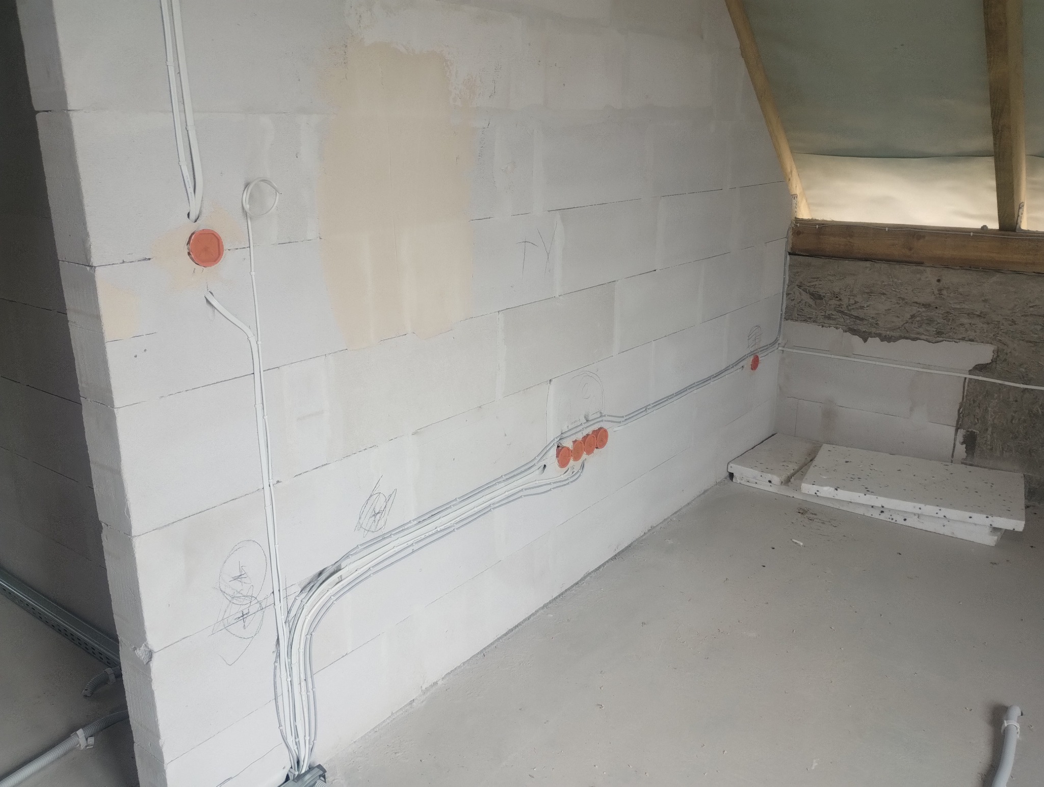 Białe ściany z instalacją elektryczną w trakcie montażu. Widoczne kable, puszki elektryczne i fragment poddasza z drewnianą konstrukcją. Szare podłoże.