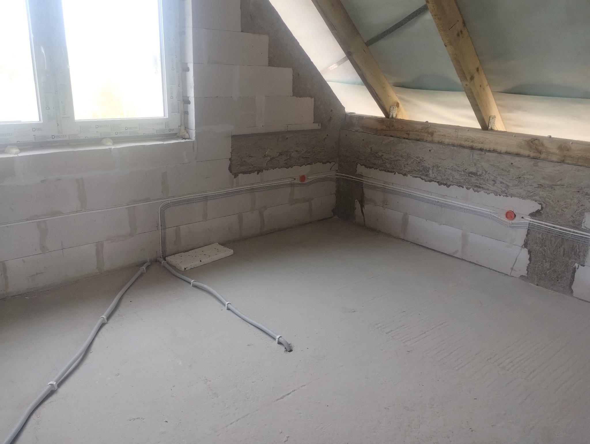 Surowe wnętrze poddasza z oknem, widoczną instalacją elektryczną w peszlach i elementami konstrukcji dachu. Ściany z betonu komórkowego.