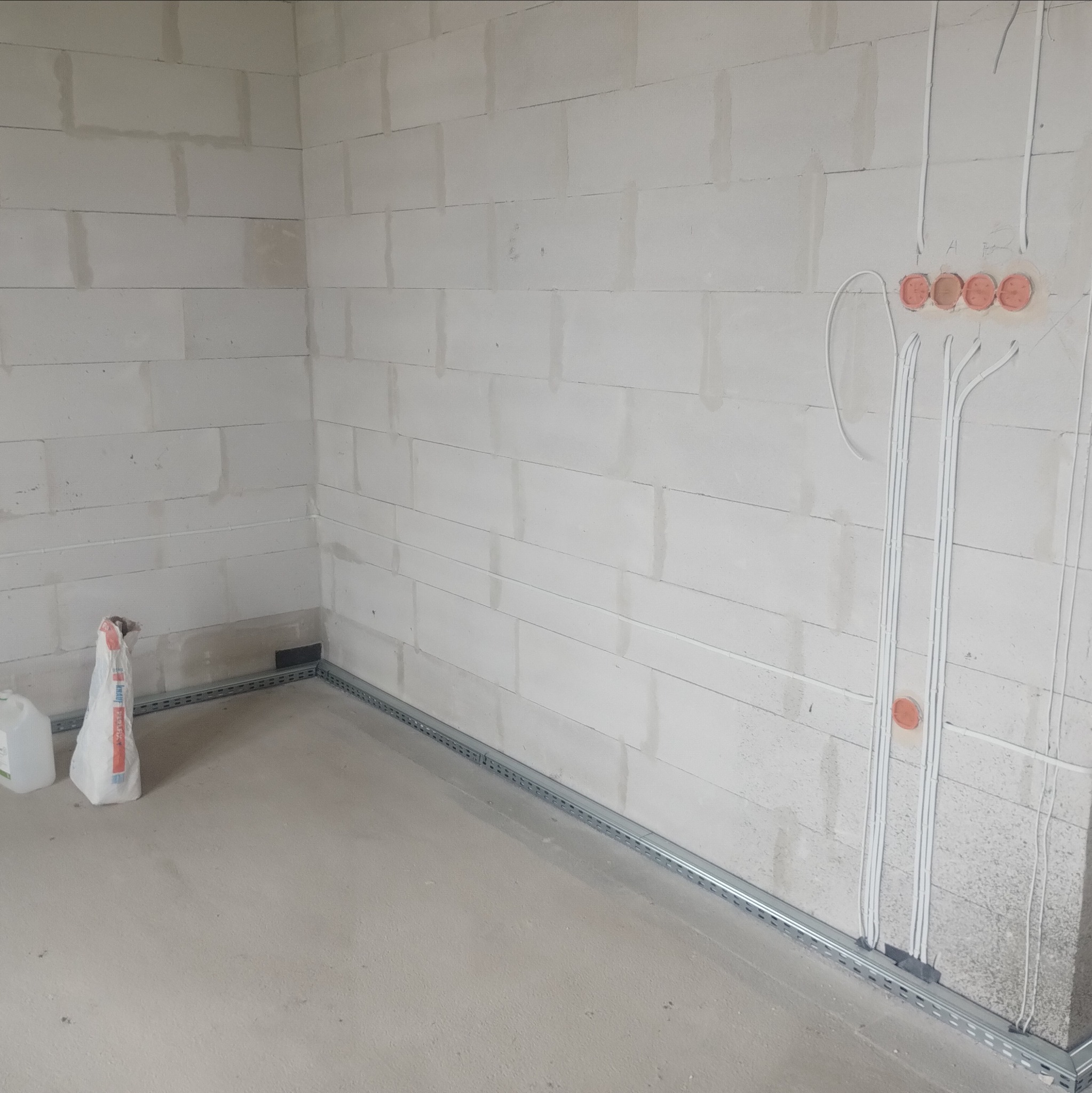 Surowy mur z instalacją elektryczną: kable w peszlach prowadzone wzdłuż ściany, puszki instalacyjne w kolorze pomarańczowym, listwy przypodłogowe. Etap prac.