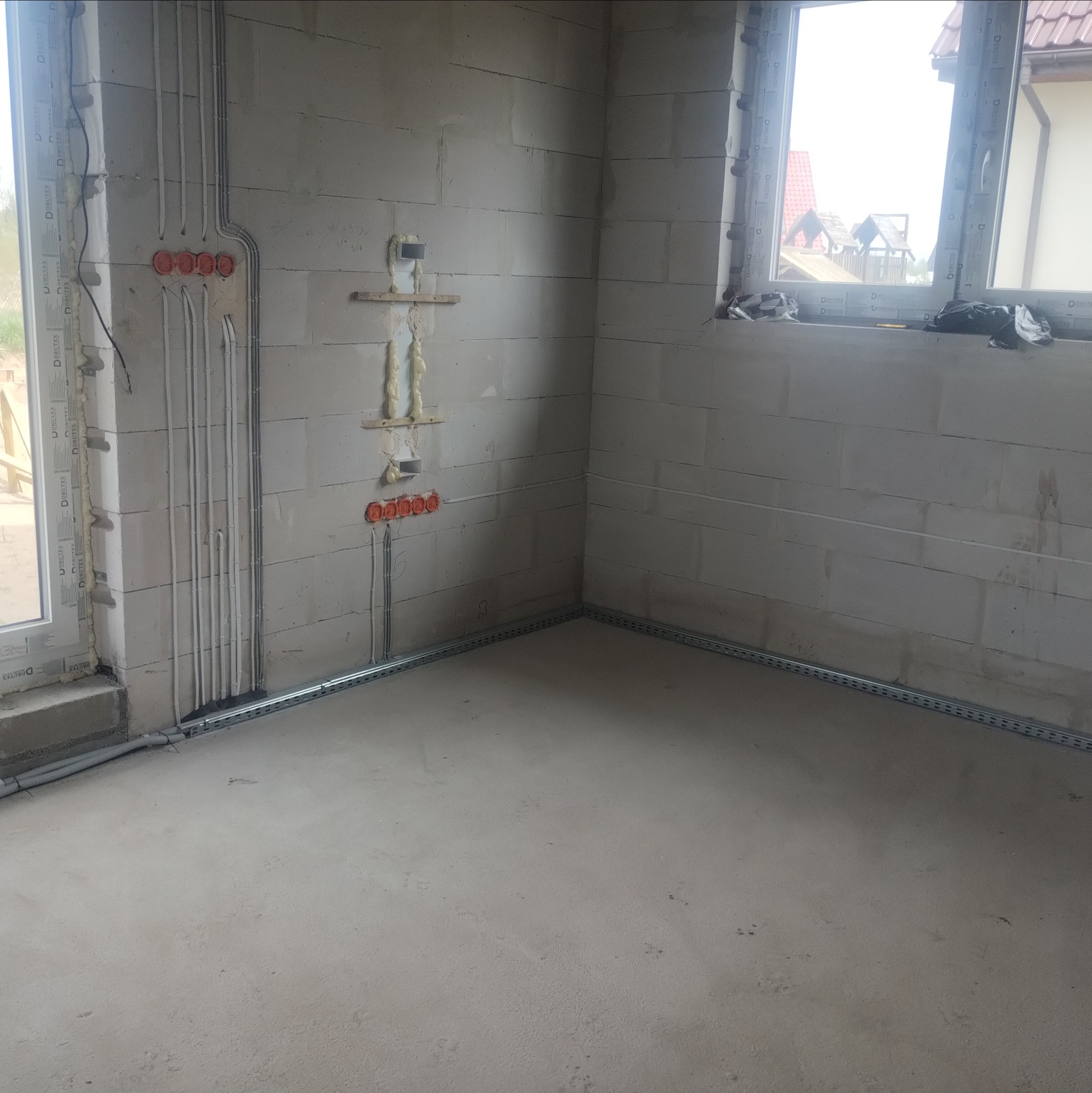 Surowy stan: wnętrze z instalacją elektryczną w peszlach i puszkami podtynkowymi, listwa przypodłogowa. Widoczne okno i ściany z betonu komórkowego.